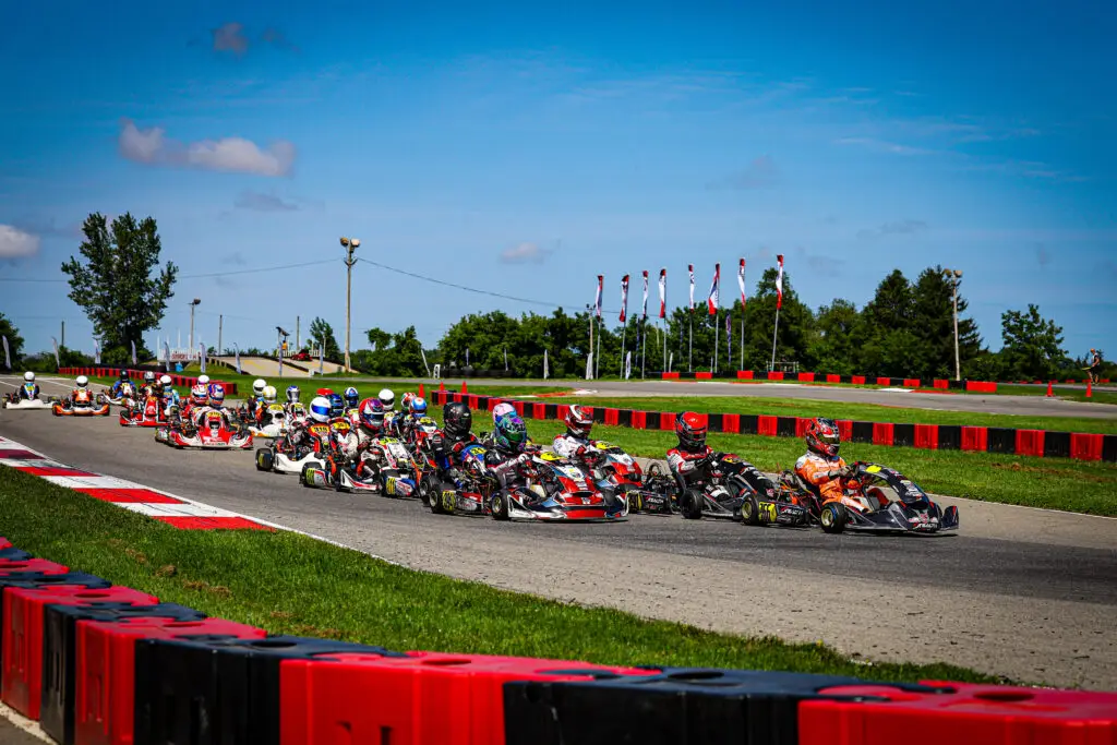 Group of go-kart racers in colorful suits navigate a curve on an outdoor track, lined with red barriers under a bright blue sky.