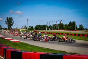 Group of go-kart racers in colorful suits navigate a curve on an outdoor track, lined with red barriers under a bright blue sky.