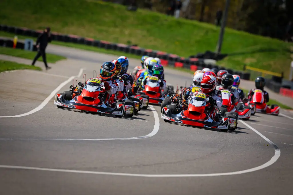 Group of go-karts with drivers leaning into a sharp turn on a race track; colorful helmets and suits visible under bright daylight.