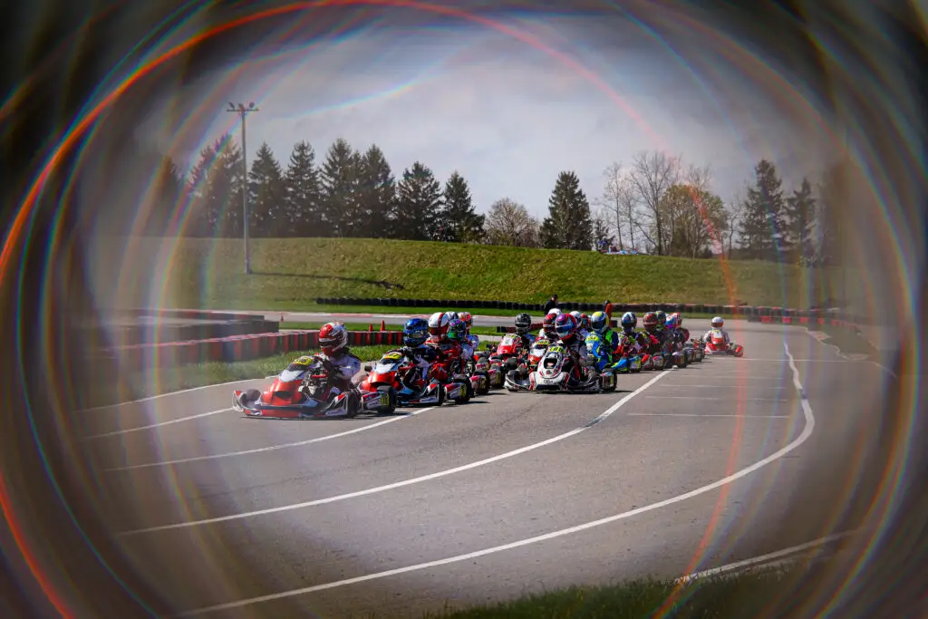 Group of go-kart racers speeding around a track with barriers and grassy infield, framed by a rainbow-colored lens flare.