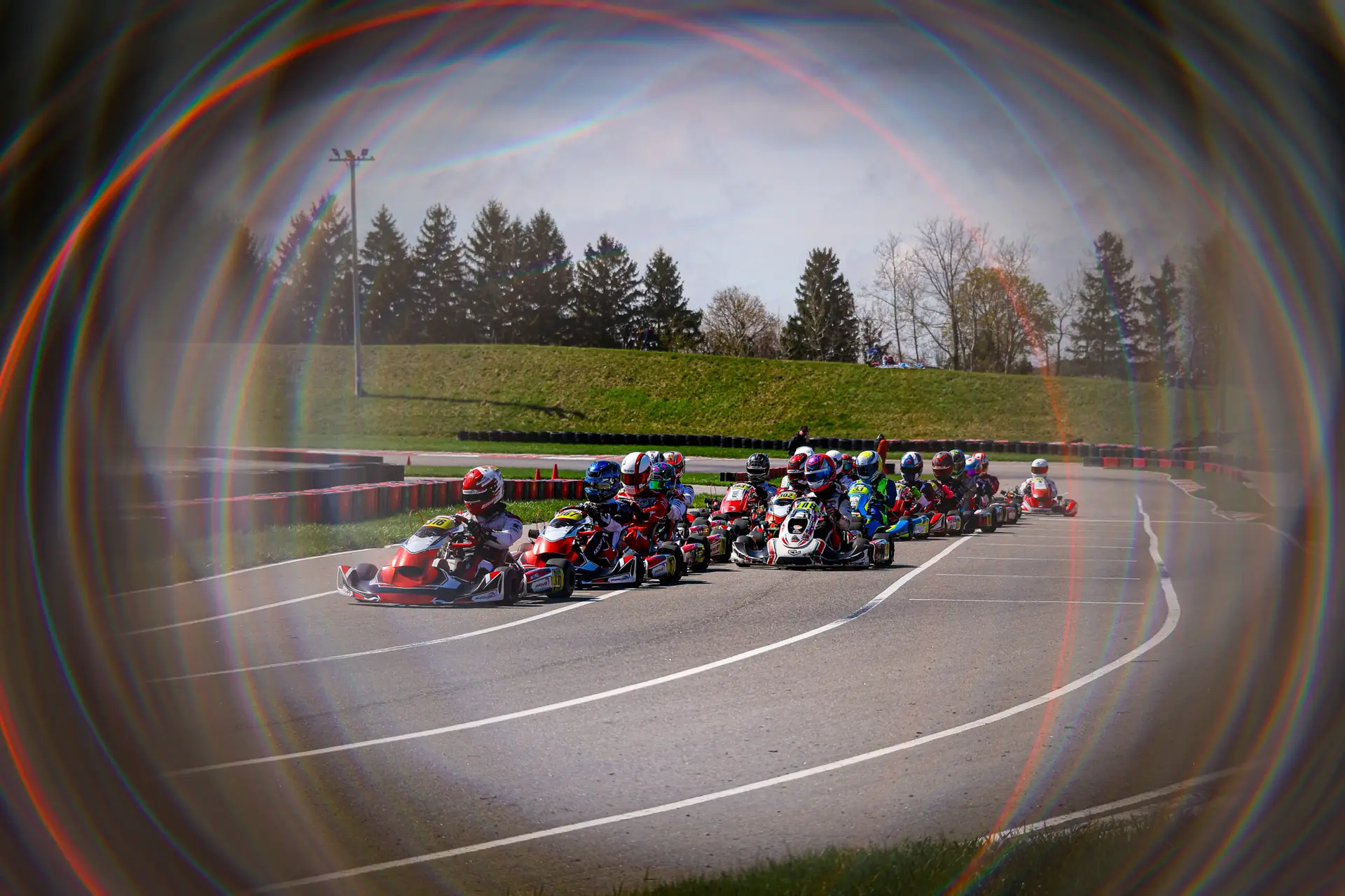 Group of go-kart racers speeding around a track with barriers and grassy infield, framed by a rainbow-colored lens flare.