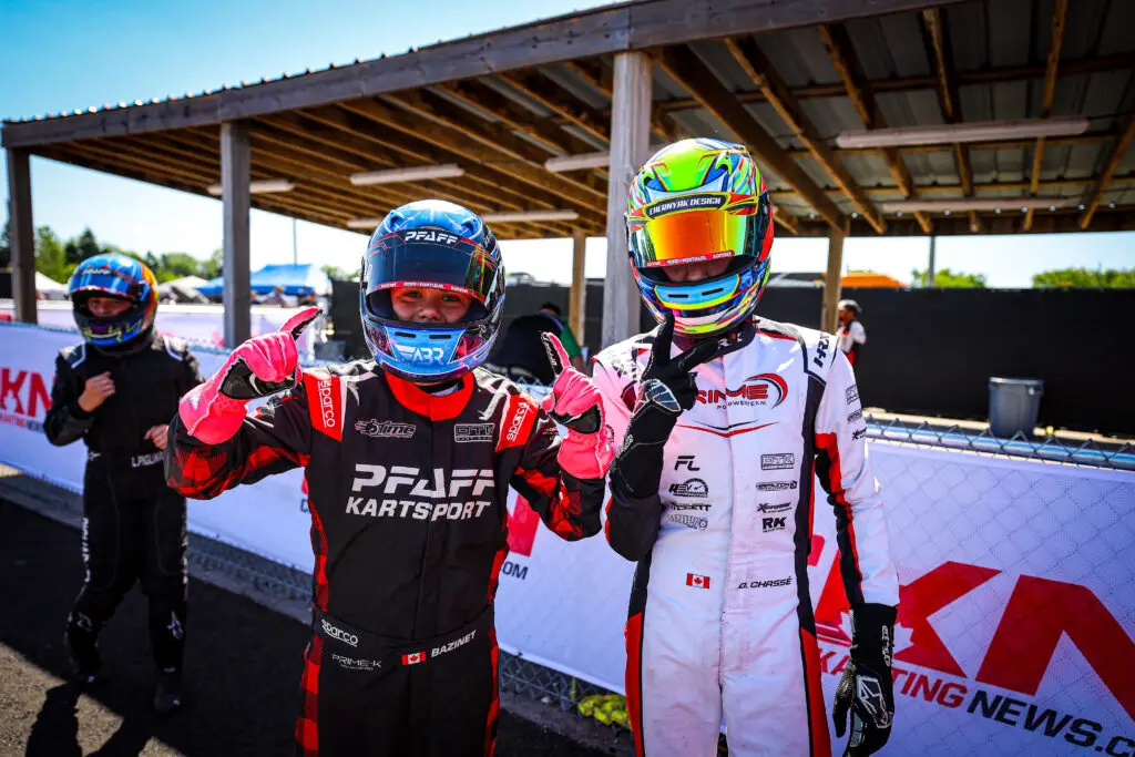 Two kart racers in colorful helmets and racing suits pose and give thumbs up at the pit area.