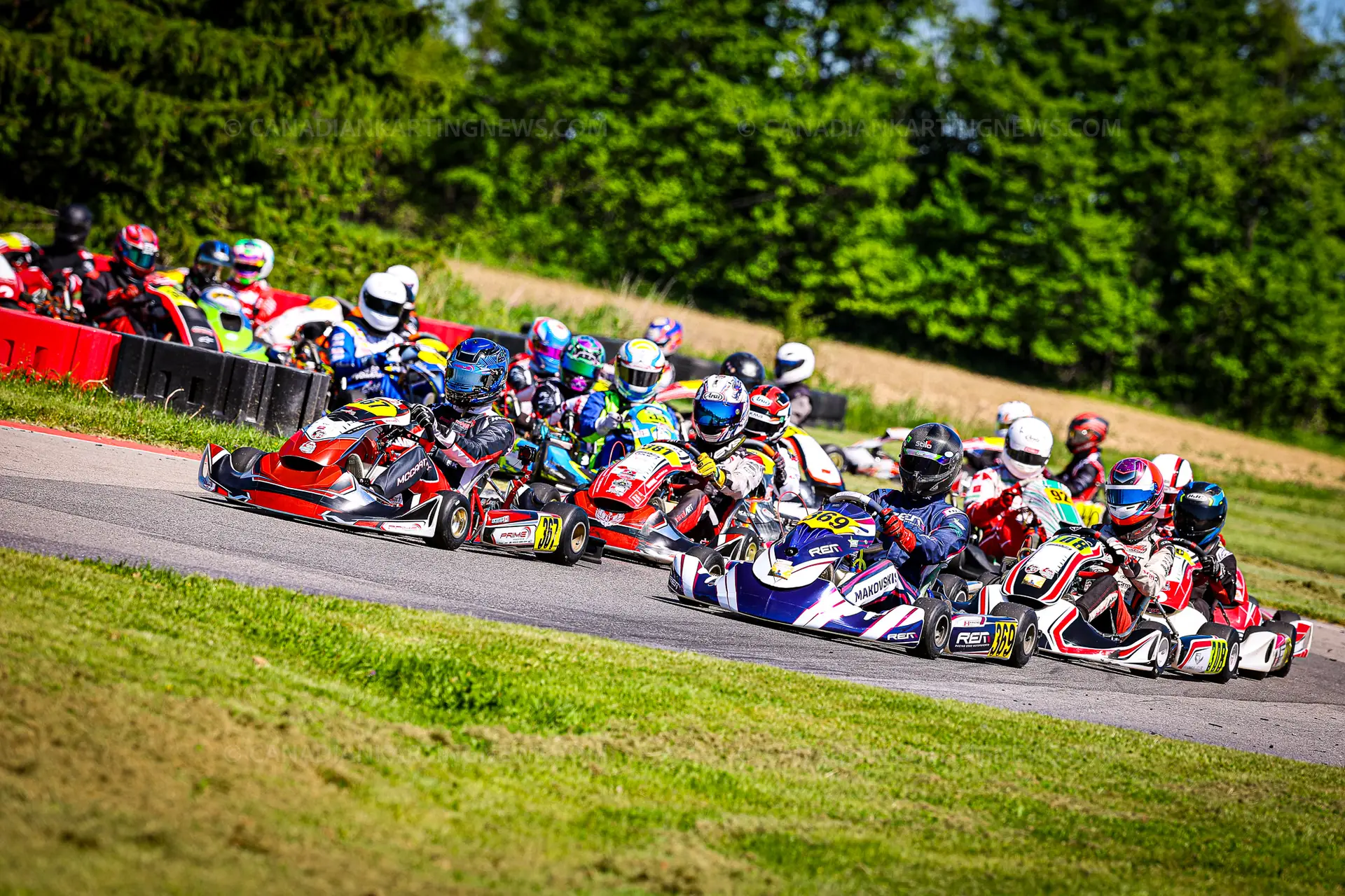 367OWENMCCARTHY369MAJORMAKOVSKIS26RMCONTR1CKN8922 Hamilton Karting Complex Go-kart racers lean into a sharp turn on a track, colorful helmets and suits creating a rainbow of tones against a green infield and tires along the barrier edge behind them.