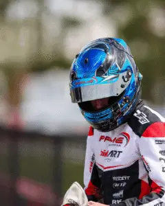 6 Hamilton Karting Complex Hamilton Karting Complex Motorsport racer wearing a blue patterned helmet and white-red racing suit with sponsor logos, preparing for a race.