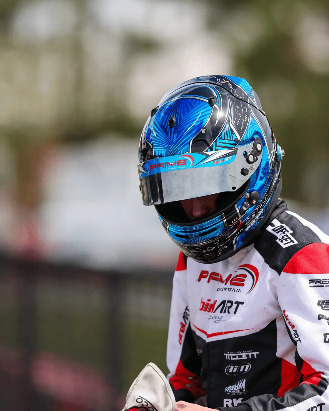 6 Hamilton Karting Complex Motorsport racer wearing a blue patterned helmet and white-red racing suit with sponsor logos, preparing for a race.