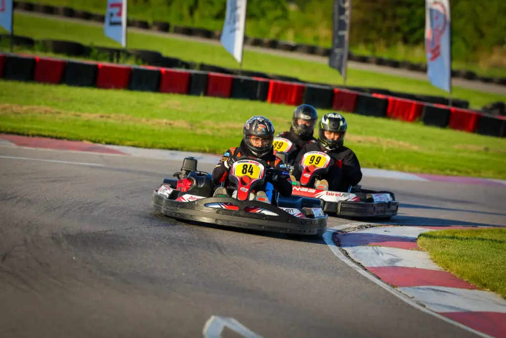 Go-k kart racers in helmets navigate a curve on an outdoor track, close formation behind each other, numbered 84 and 99 to the foreground.