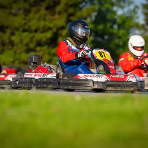 Three go-kart racers in helmets speeding around a track; center kart number 87 in blue-red gear, others numbered 93 and 94 in red suits.