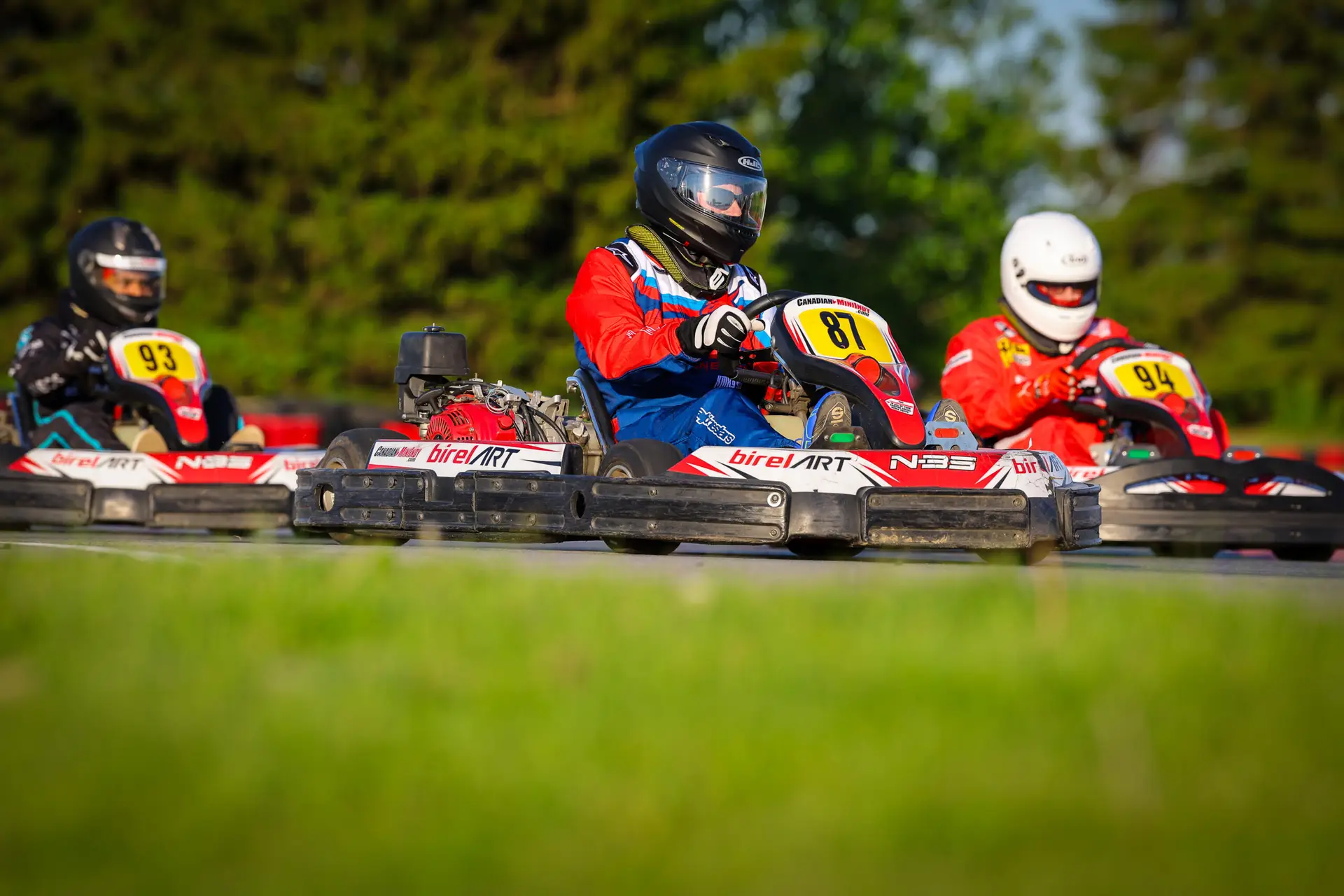 CKN1075 Hamilton Karting Complex Three go-kart racers in helmets speeding around a track; center kart number 87 in blue-red gear, others numbered 93 and 94 in red suits.