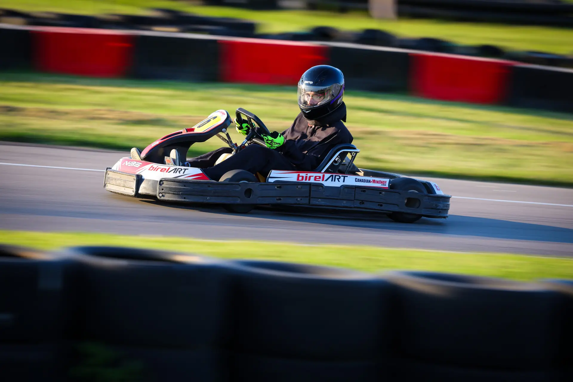 Go-kart racer in black suit and helmet speeds around a track with red and blue barriers in the background.