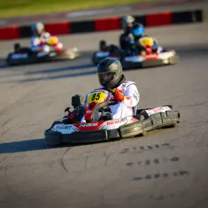 Go-kart racer in a white suit and helmet leads a race on a sunny track with others blurred in the background.