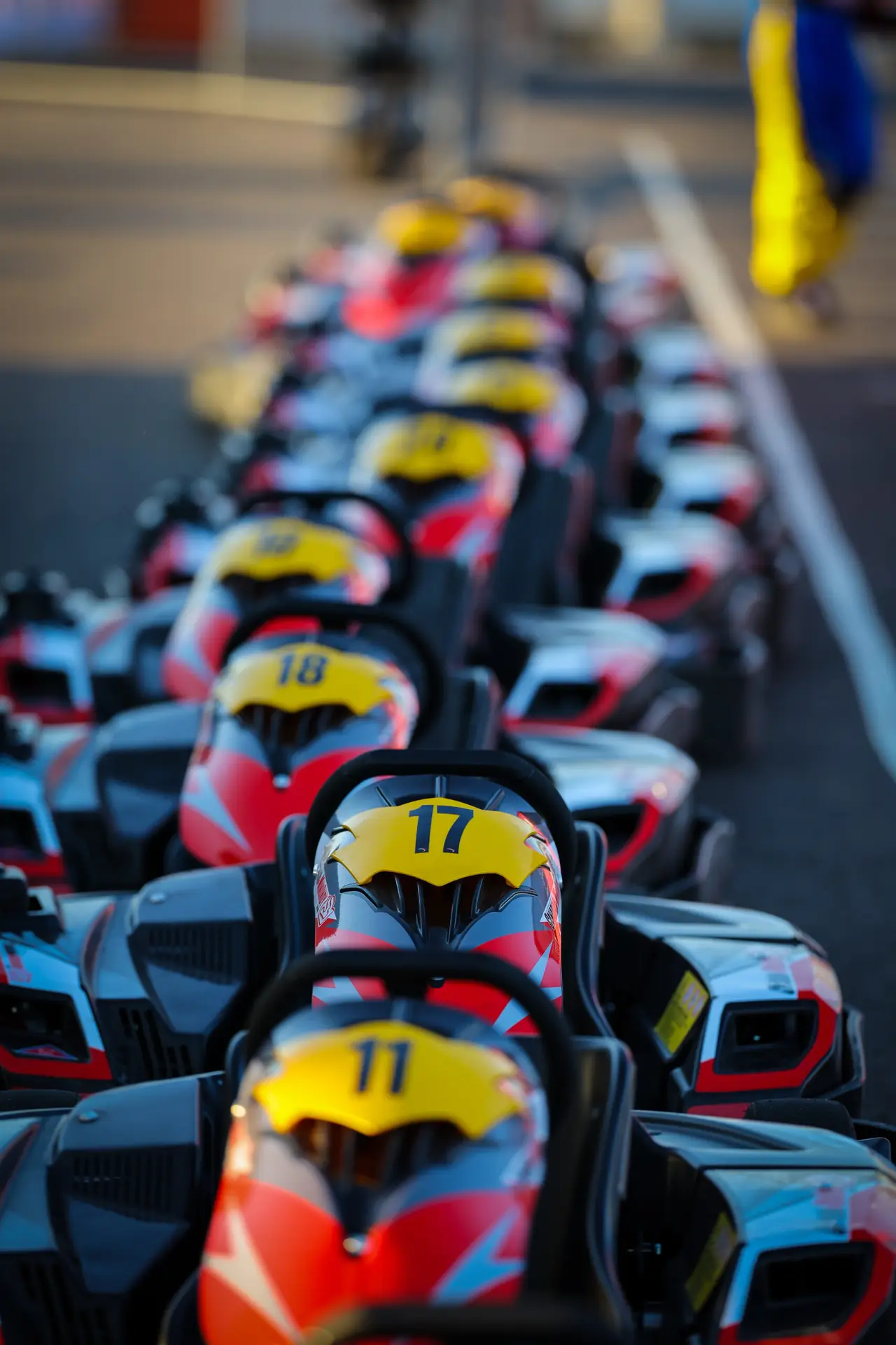 Row of go-karts with yellow helmets lined up on a karting track, numbers 17 and 11 visible (blurred).