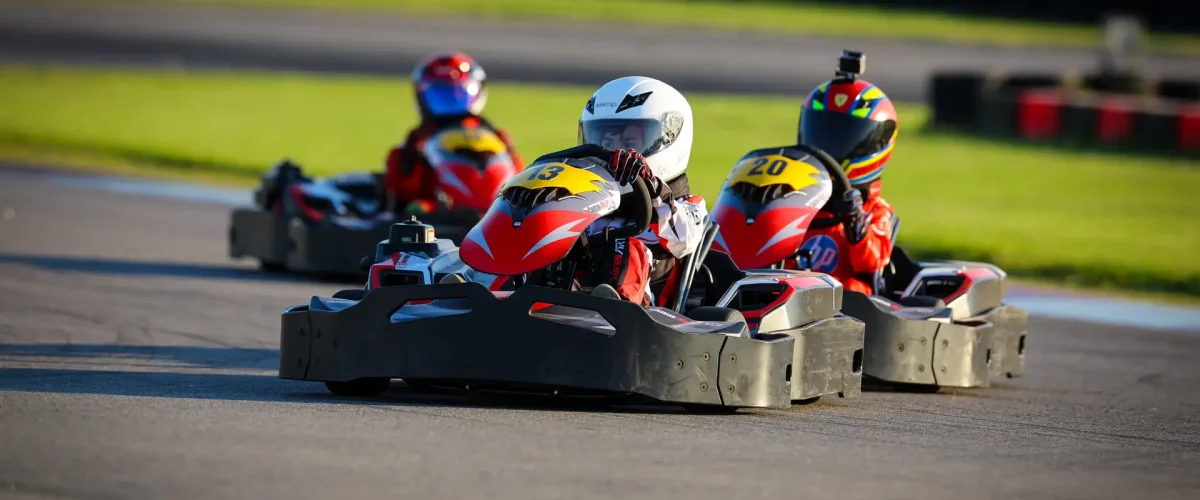 Go-kart racers in colorful helmets speed around a sunny outdoor track in a close race.