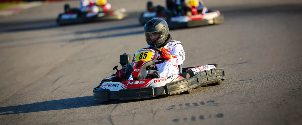 Go-kart racer in a white suit and helmet leads a race on a sunny track with others blurred in the background.